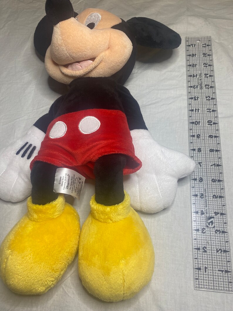 Puede incluir: Un juguete de peluche de Mickey Mouse con pelaje negro, pantalones cortos rojos con lunares blancos y zapatos amarillos. El juguete mide aproximadamente 50 cm de alto.