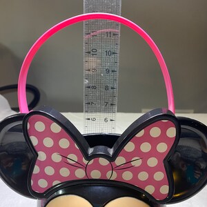 Puede incluir: Un cubo de pl&aacute;stico negro de Minnie Mouse con un lazo rosa de lunares blancos y un asa rosa. El cubo mide aproximadamente 15 cm de alto.