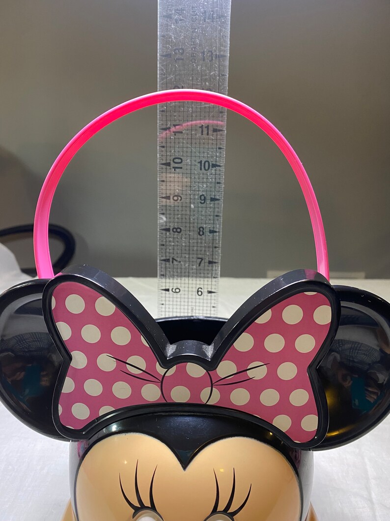 Puede incluir: Un cubo de pl&aacute;stico negro de Minnie Mouse con un lazo rosa de lunares blancos y un asa rosa. El cubo mide aproximadamente 15 cm de alto.