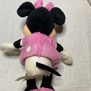 Puede incluir: Un juguete de peluche Minnie Mouse rosa y negro con un vestido blanco de lunares y un lazo rosa. El juguete est&aacute; sentado sobre una superficie blanca.