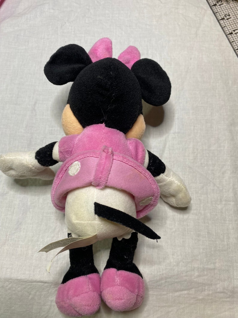 Puede incluir: Un juguete de peluche Minnie Mouse rosa y negro con un vestido blanco de lunares y un lazo rosa. El juguete est&aacute; sentado sobre una superficie blanca.