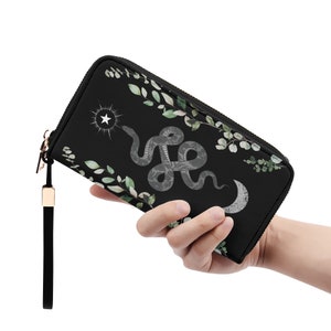 May include: Black wallet with a snake, star, and moon design. The wallet has a zip closure, a black wrist strap, and a gold-coloured metal zip pull. The snake is grey with a floral pattern, and the design includes green leaves.
