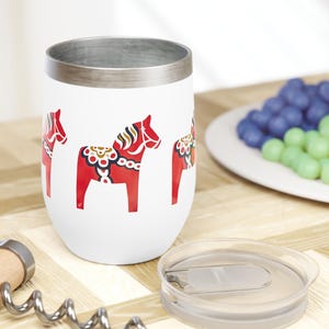 May include: A white insulated wine tumbler with a silver rim and a red Dala horse design. A wooden corkscrew and clear lid are next to the tumbler. A plate of colorful candies is in the background.