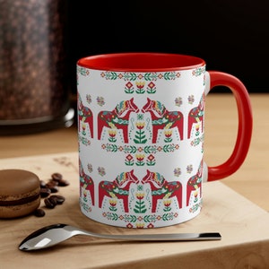 May include: A white ceramic mug with a red rim and handle. The mug features a repeating pattern of red, yellow, and green Dala horses with floral accents.