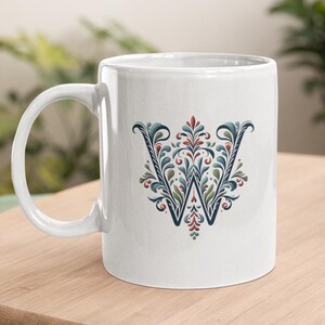 May include: White ceramic mug with a decorative floral "W" design in blue, green, and red. The mug has a curved handle and is placed on a wooden surface. The mug is suitable for hot or cold beverages.