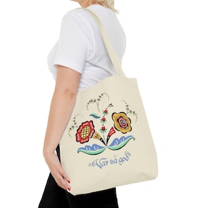 May include: Cream-colored tote bag featuring a colorful floral design with red, yellow, blue, and green accents. The bag has a shoulder strap and the text "Var så god" in blue. The bag is made of a sturdy fabric.