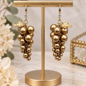 May include: A pair of gold-toned dangle earrings shaped like grape clusters, featuring multiple metallic gold spheres, a small gold leaf accent, and a single clear crystal set at the top of each earring, suspended from a gold-toned T-bar display stand.
