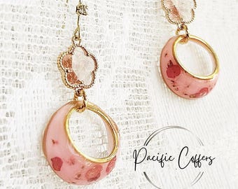 May include: Dangle earrings with a floral design. The earrings have gold-coloured metal with pink, oval-shaped pendants. The pendants have a clear centre and are decorated with red accents. The earrings are on a white lace background.