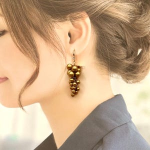 May include: A drop earring featuring a cluster of metallic bronze-coloured spherical beads arranged in a grape-like formation. The beads are suspended from a small gold-toned metal hook with a tiny clear crystal accent at the top of the cluster.