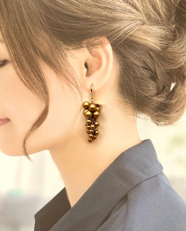 May include: A drop earring featuring a cluster of metallic bronze-coloured spherical beads arranged in a grape-like formation. The beads are suspended from a small gold-toned metal hook with a tiny clear crystal accent at the top of the cluster.
