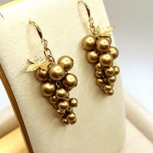 May include: A pair of gold-toned drop earrings shaped like clusters of grapes. Each earring features multiple round, metallic gold beads arranged in a bunch, accented with a textured gold leaf at the top and a small clear stone on the hook wire.