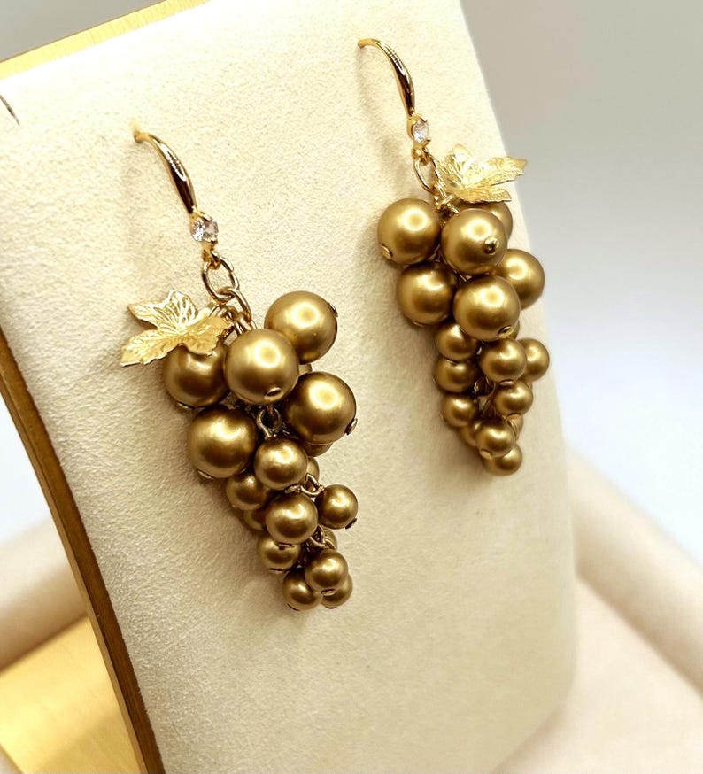 May include: A pair of gold-toned drop earrings shaped like clusters of grapes. Each earring features multiple round, metallic gold beads arranged in a bunch, accented with a textured gold leaf at the top and a small clear stone on the hook wire.
