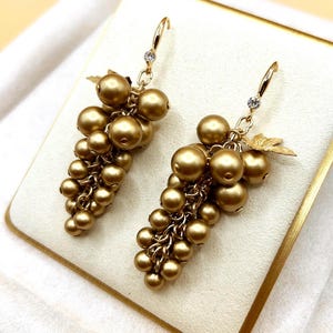 May include: A pair of gold-toned drop earrings shaped like clusters of grapes. Each earring features numerous round, matte gold beads suspended from a gold-coloured hook, accented with a small clear crystal and a metallic leaf detail at the top.