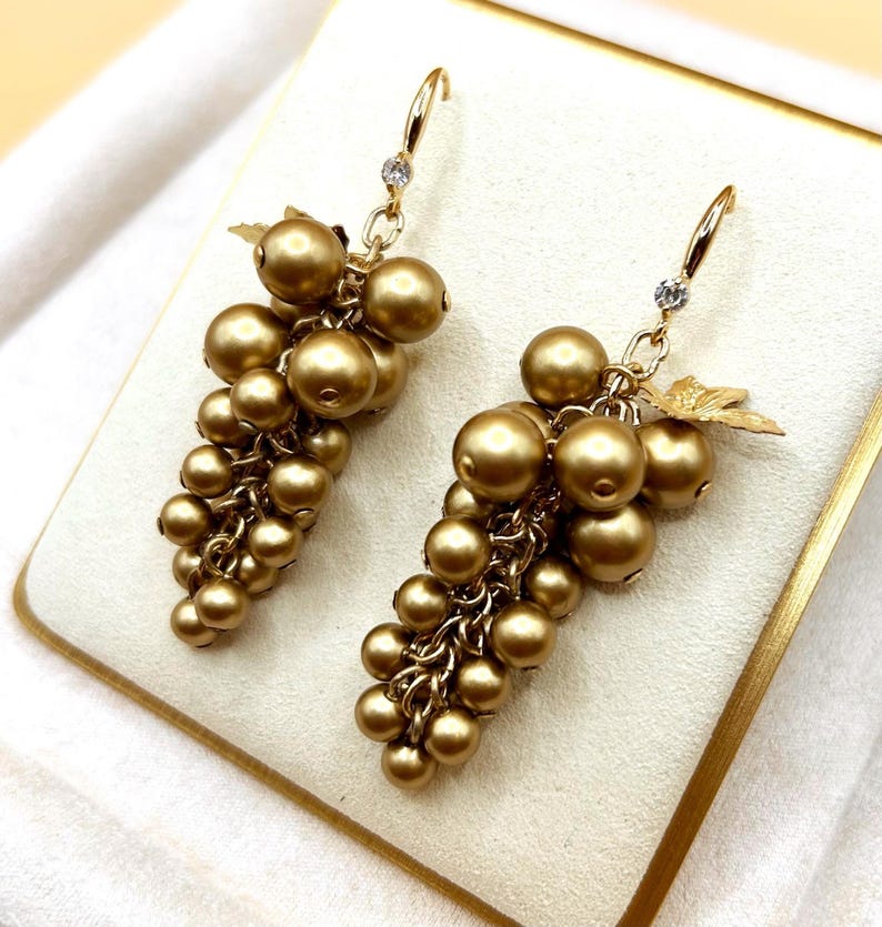 May include: A pair of gold-toned drop earrings shaped like clusters of grapes. Each earring features numerous round, matte gold beads suspended from a gold-coloured hook, accented with a small clear crystal and a metallic leaf detail at the top.