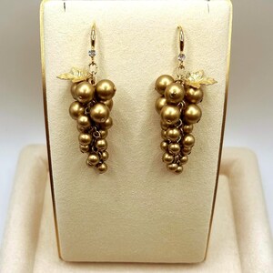 May include: A pair of drop earrings featuring clusters of metallic gold spherical beads arranged to resemble bunches of grapes. Each earring includes a small gold leaf accent at the top and a single clear stone set into the gold hook wire mechanism.