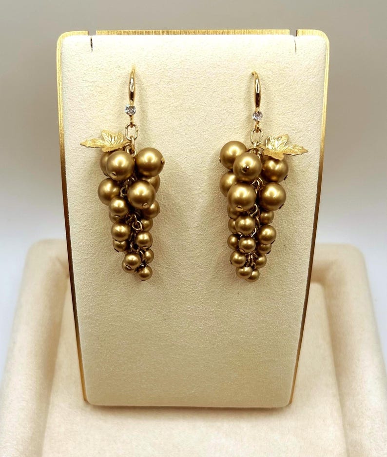 May include: A pair of drop earrings featuring clusters of metallic gold spherical beads arranged to resemble bunches of grapes. Each earring includes a small gold leaf accent at the top and a single clear stone set into the gold hook wire mechanism.