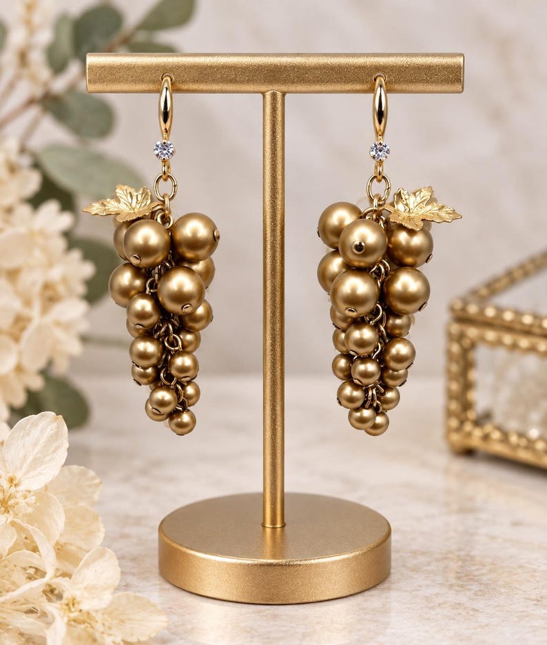 May include: A pair of gold-toned dangle earrings shaped like grape clusters, featuring multiple metallic gold spheres, a small gold leaf accent, and a single clear crystal set at the top of each earring, suspended from a gold-toned T-bar display stand.