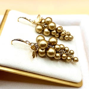 May include: A pair of drop earrings featuring clusters of metallic gold spherical beads arranged to resemble grapes. Each earring includes a small gold leaf accent near the top and a curved hook wire with a tiny clear stone at the connection point.