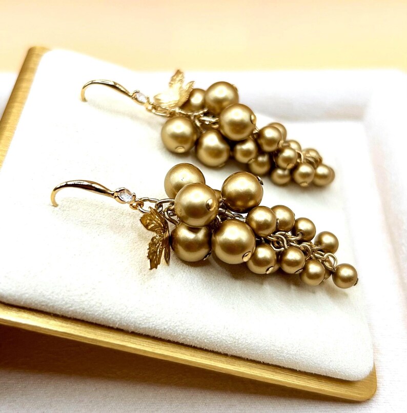 May include: A pair of drop earrings featuring clusters of metallic gold spherical beads arranged to resemble grapes. Each earring includes a small gold leaf accent near the top and a curved hook wire with a tiny clear stone at the connection point.