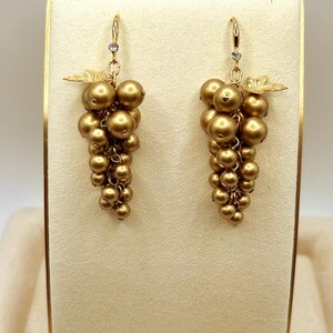 May include: A pair of gold-toned drop earrings shaped like clusters of grapes. Each earring features multiple round, matte gold beads suspended from a hook, accented with a small clear crystal at the top and a delicate gold leaf detail near the hook.