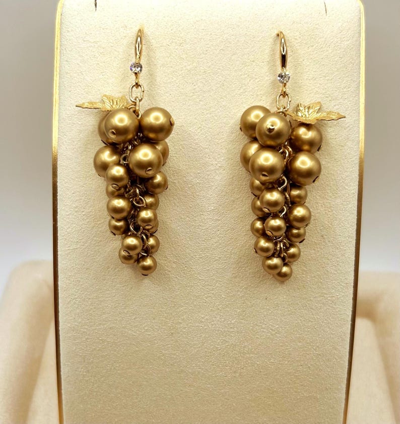 May include: A pair of gold-toned drop earrings shaped like clusters of grapes. Each earring features multiple round, matte gold beads suspended from a hook, accented with a small clear crystal at the top and a delicate gold leaf detail near the hook.