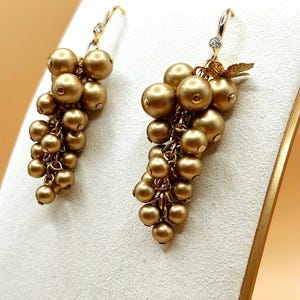 May include: A pair of drop earrings featuring clusters of matte gold-toned spherical beads arranged to resemble bunches of grapes. Each earring is suspended from a gold-toned hook with a small, clear, faceted stone accent at the top.