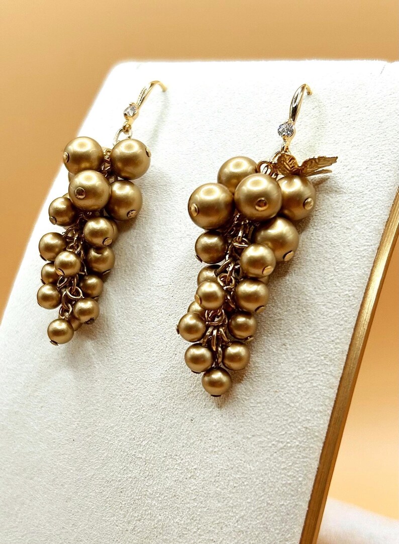 May include: A pair of drop earrings featuring clusters of matte gold-toned spherical beads arranged to resemble bunches of grapes. Each earring is suspended from a gold-toned hook with a small, clear, faceted stone accent at the top.