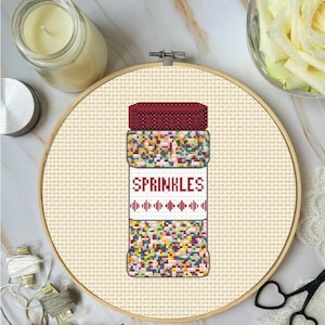May include: Cross-stitch design of a jar of sprinkles with a burgundy lid and a white label that reads "SPRINKLES". The jar is filled with colorful sprinkles. The design is set in a wooden embroidery hoop.