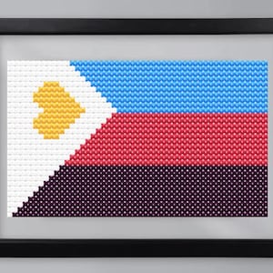 May include: Framed cross-stitch artwork depicting the flag of the Philippines. The flag features a white triangle with a yellow sun, a blue stripe, a red stripe, and a dark purple stripe. The artwork is framed in a black frame.