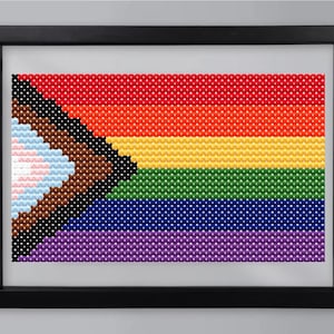 May include: Framed cross-stitch artwork featuring the Progress Pride flag. The flag design includes the traditional rainbow stripes, along with black, brown, light blue, pink, and white chevrons. The artwork is set in a black frame.