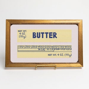 May include: A framed cross-stitch design of a butter stick label. The label reads "NET WT. 4 OZ. (113g)" and "BUTTER". The label also includes a measurement guide for tablespoons and cups.