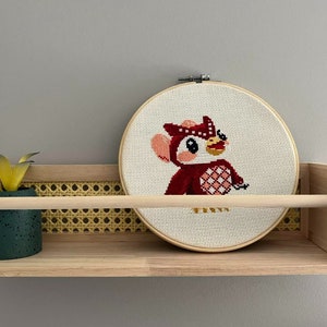 May include: A cross-stitch hoop art featuring a cartoon owl in shades of red, pink, and white. The artwork is displayed on a wooden shelf with a small potted plant. The shelf has a natural wood finish and a woven cane detail.