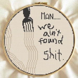 May include: A white embroidery hoop with black text that reads "Man... We ain't found shit."