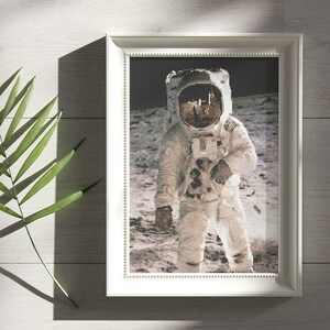 Op de afbeelding: Een ingelijste zwart-witfoto van een astronaut die op de maan staat. De astronaut draagt een wit ruimtepak met een helm en kijkt naar het maanoppervlak.