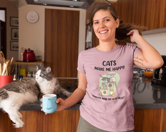 Puede incluir: Una mujer lleva una camiseta rosa con el texto "Los gatos me hacen feliz, los humanos me dan dolor de cabeza" y un gráfico de gatos en un árbol para gatos. Un gato está sentado en una encimera junto a una taza azul.