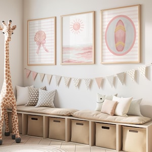 Könnte beinhalten: Kinderzimmer mit drei gerahmten Aquarelldrucken mit einer Muschel, einer Sonne über dem Meer und einem Surfbrett. Ein Giraffenspielzeug, Kissen und eine Sitzbank mit Banner vervollständigen die Einrichtung.