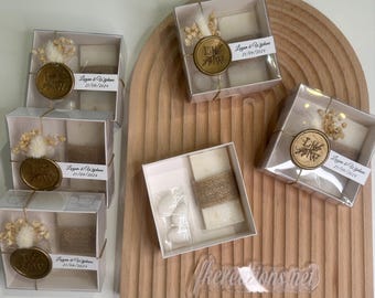 May include: A set of six small, white gift boxes with clear lids. Each box contains a small white square, a sprig of dried flowers, and a gold wax seal with the initials "L & W" and the date "21/09/2024". The boxes are arranged on a wooden surface.