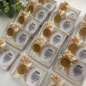 Puede incluir: Una colección de pequeñas cajas transparentes llenas de platos decorativos redondos blancos con caligrafía árabe. Cada caja contiene un sello de cera dorado, flores secas y purpurina dorada. Los platos muestran el texto "Make a Wish" y "Eid Mubarak".