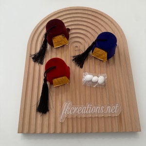 Peut inclure: Trois petits fez en velours rouge, bordeaux et bleu avec des garnitures dorées et des glands noirs. Les fez sont disposés sur un plateau en bois avec un sachet de bonbons blanc.