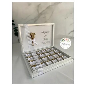 Peut inclure: Boîte cadeau blanche avec un ruban blanc et un petit bouquet de fleurs séchées. Le couvercle de la boîte porte l'inscription "Baptême de Nelia 14.10.2023". La boîte est remplie de petites faveurs en chocolat blanc.