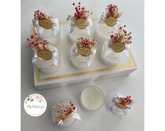 May include: Set of eight small glass candle jars with white lids and white bows. Each jar has a dried flower arrangement and a gold sticker with a name and date. The jars are arranged on a white box with gold trim.