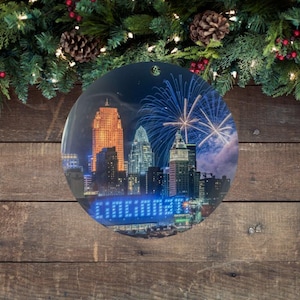 May include: A round Christmas ornament featuring a nighttime cityscape with fireworks. The ornament has the word "Cincinnati" in blue lights. The background is a wooden surface with evergreen branches, pinecones, and red berries.