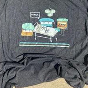 May include: A dark gray t-shirt with a cartoon illustration of four characters. The characters are a green leafy vegetable, a brown rectangle with a face, a blue rectangle with a face, and a pencil with a face. The pencil is saying "Pencil" in a speech bubble. The characters are standing around a hospital bed with a white sheet and a medical symbol on it.
