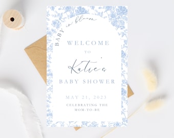 May include: A baby shower welcome sign with a floral design in light blue. The sign reads "Welcome to Katie's Baby Shower" with the date May 21, 2023. The sign is on a white background with a brown envelope.