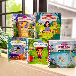 May include: Five colorful children's books with lift-the-flap features. The books feature classic fairy tales such as Rapunzel, Snow White, Sleeping Beauty, Pinocchio, and The Ugly Duckling. The books are displayed on a windowsill with a green plant in the foreground.