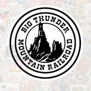 May include: A circular sticker with a black and white design. The text "BIG THUNDER MOUNTAIN RAILROAD" is printed around the outer edge. Inside the circle is a silhouette of a mountain range. The sticker is surrounded by other colorful stickers.