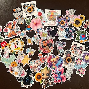 May include: A collection of colorful stickers featuring Disney characters and floral designs. The stickers include images of Mickey Mouse, Bambi, Tinkerbell, and other characters, with sunflowers, roses, and other flowers. The stickers are arranged on a dark surface.