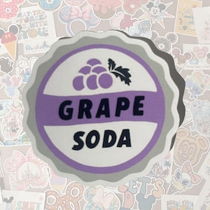 May include: A round, gray and white sticker with the words "GRAPE SODA" in black, and a purple grape illustration. The sticker has a scalloped edge and is surrounded by other colorful stickers.