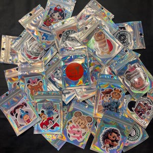 May include: A pile of holographic, resealable bags containing stickers with cartoon characters and logos. Each bag has a small hole at the top. The stickers are of various sizes and colours. The bags are arranged in a pile.