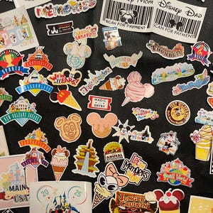 May include: A collection of colorful Disney-themed stickers and decals. The assortment includes various designs featuring Mickey Mouse, Minnie Mouse, and other characters, along with park attractions and phrases like "Family Vacation 2025" and "Memories."